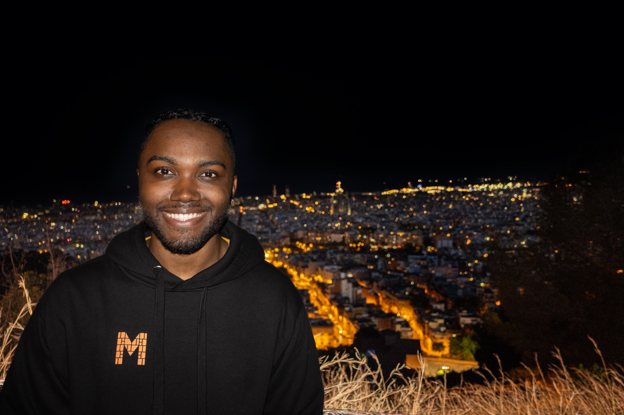 Quentin Simoneaux at Bunkers del Carmel, Barcelona, at night — mosaic-M hoodie, the lit city grid behind