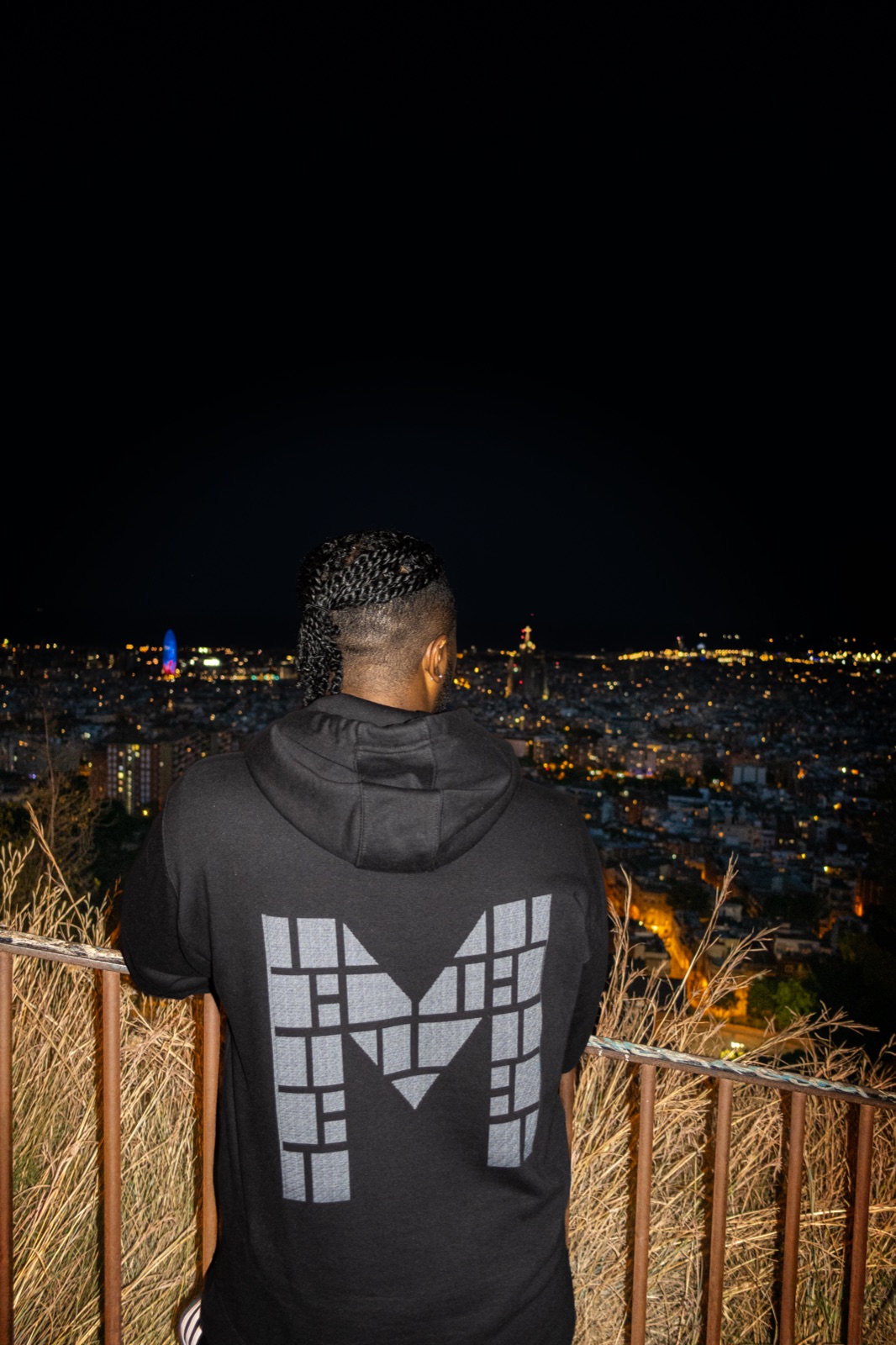The mosaic-M logo across the back of a hoodie, overlooking the lit Barcelona grid